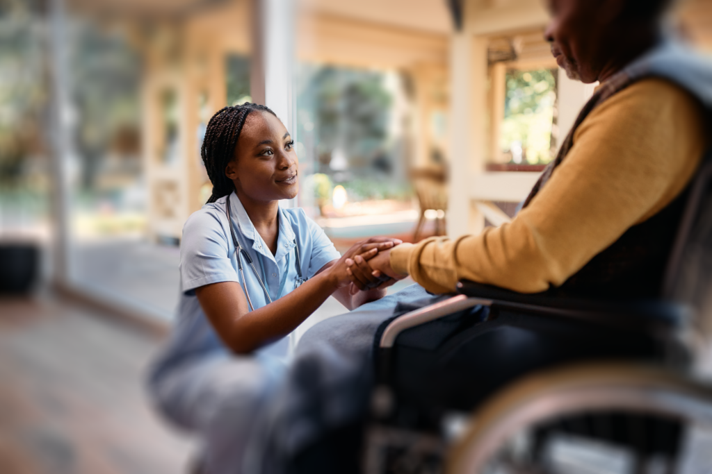A female nurse kneels in front of an elderly patient in a wheel chair holding his hand.