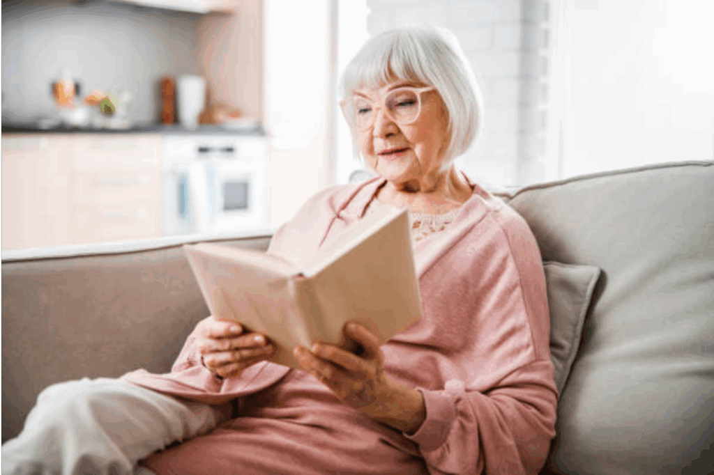 An elderly woman sits on a large chair reading a book wearing a pink sweater and glasses.