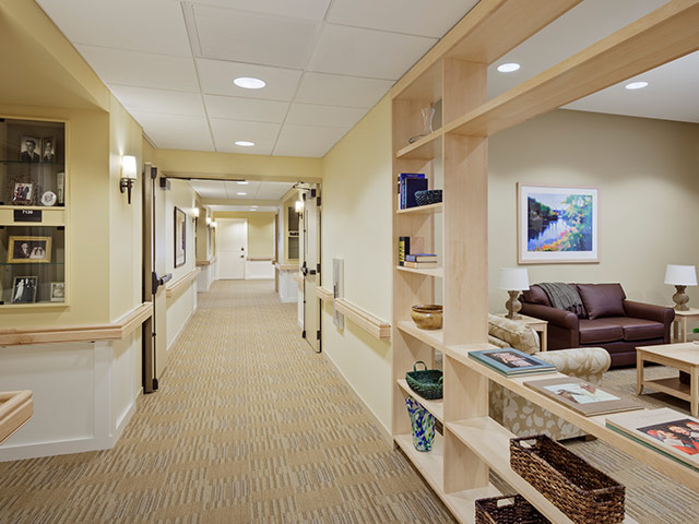 A warm, inviting hallway in a senior living community with soft lighting, built-in display shelves, and a cozy seating area with couches and artwork.