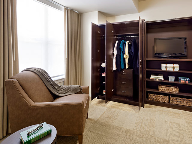 A cozy seating area in a senior living apartment featuring an upholstered armchair, natural light, and built-in wardrobe storage.
