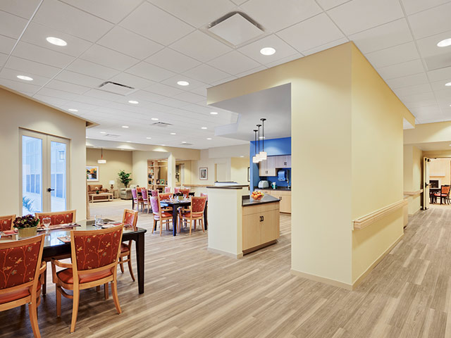 A spacious, well-lit dining area in a senior living community with neatly set tables, comfortable seating, and an open kitchen nearby.